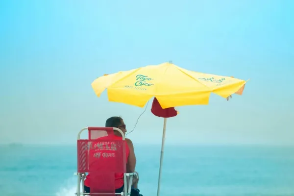 Man zit op een strandstoel op het strand, onder een gele parasol. Foto is genomen vanaf de achterkant.