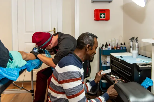 GGD medewerker controleert materiaal in een tattoo shop. Op de achtergrond is een tatoeëerder bezig.