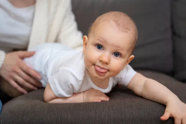 Baby ligt op de buik op de bank en kijkt met grote ogen en tong uit de mond naar de camera. De baby draagt een witte romper. Op de achtergrond is te zien dat een volwassen vrouw het kindje vasthoudt zodat het niet van de bank kan vallen.